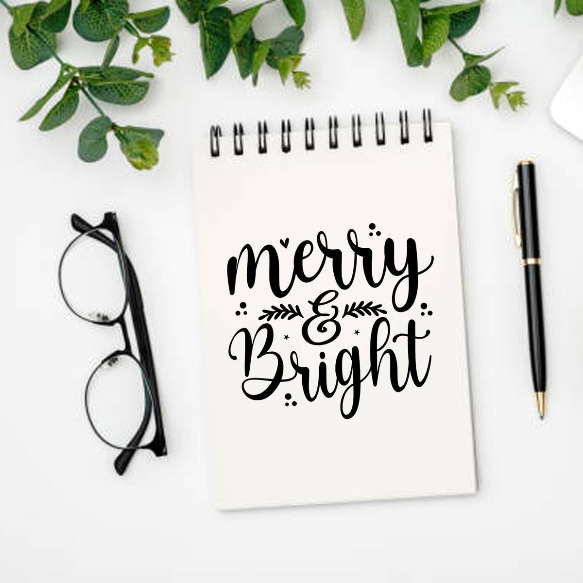 Holiday stamp and seal design featuring black merry & bright lettering on a white spiral notebook, with pen, glasses, and greenery.