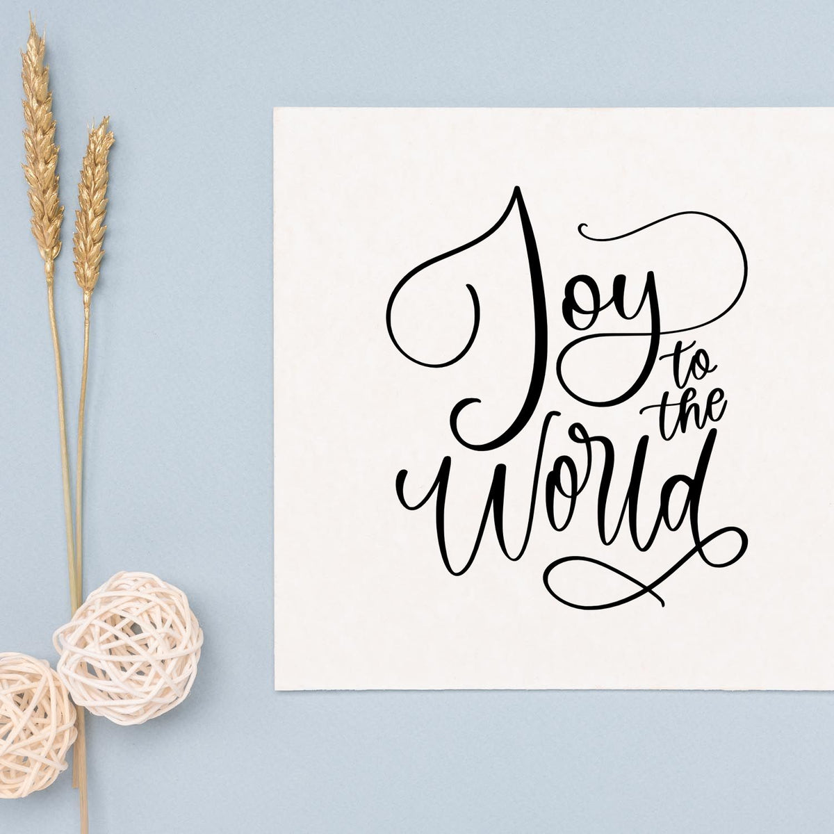 Black Joy to the world stamp/seal on white card, with gold wheat stalks and white wicker accents on pale blue background.