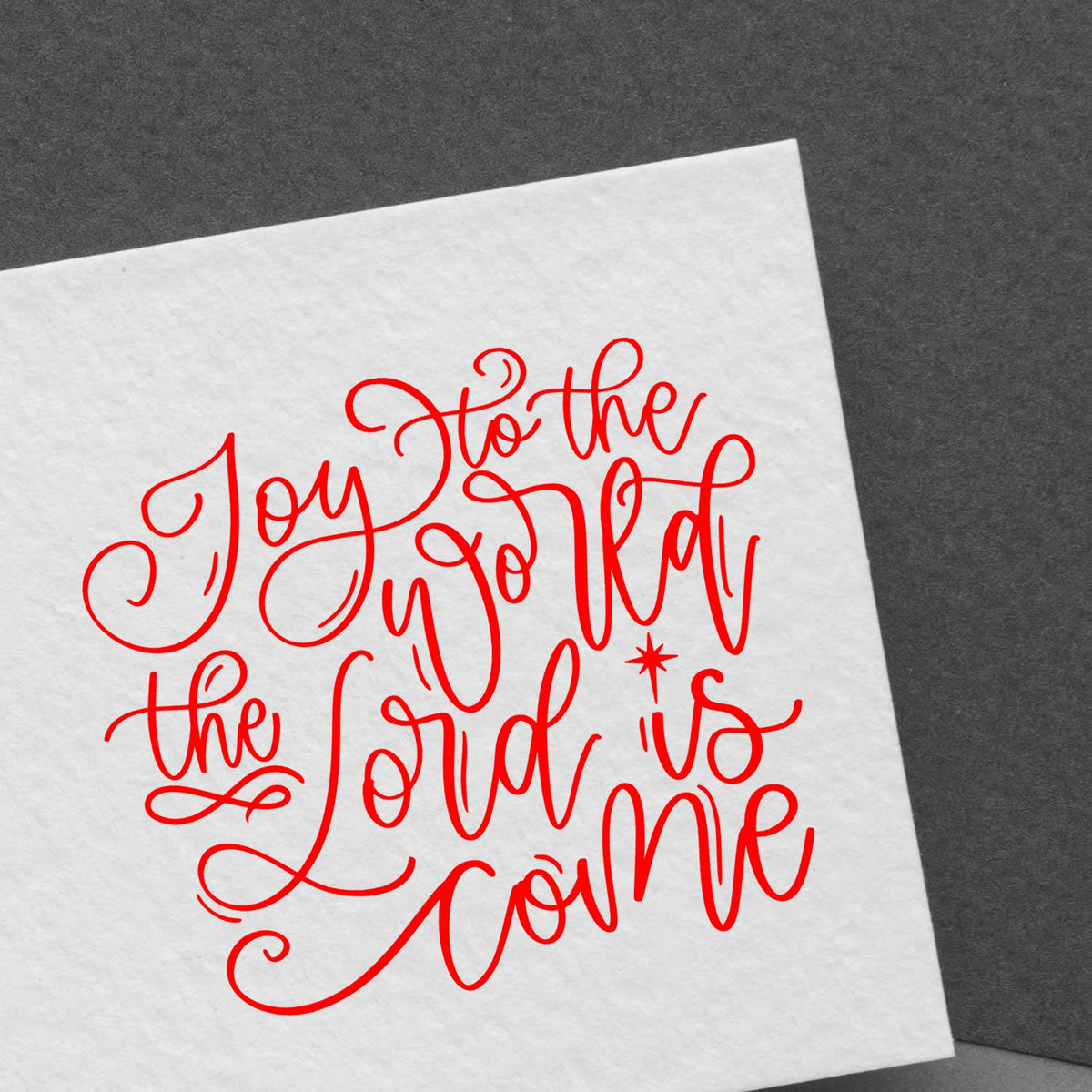 Red decorative stamp design on white paper with Joy to the world the Lord is come in script, on dark gray background.