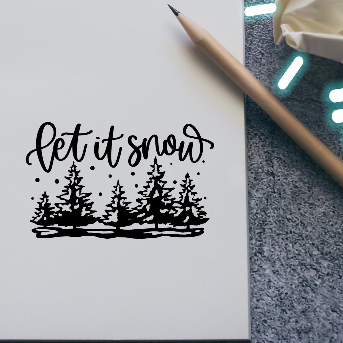 Black "let it snow" stamp seal with pine trees and snow dots on white paper, beside a pencil on gray background.