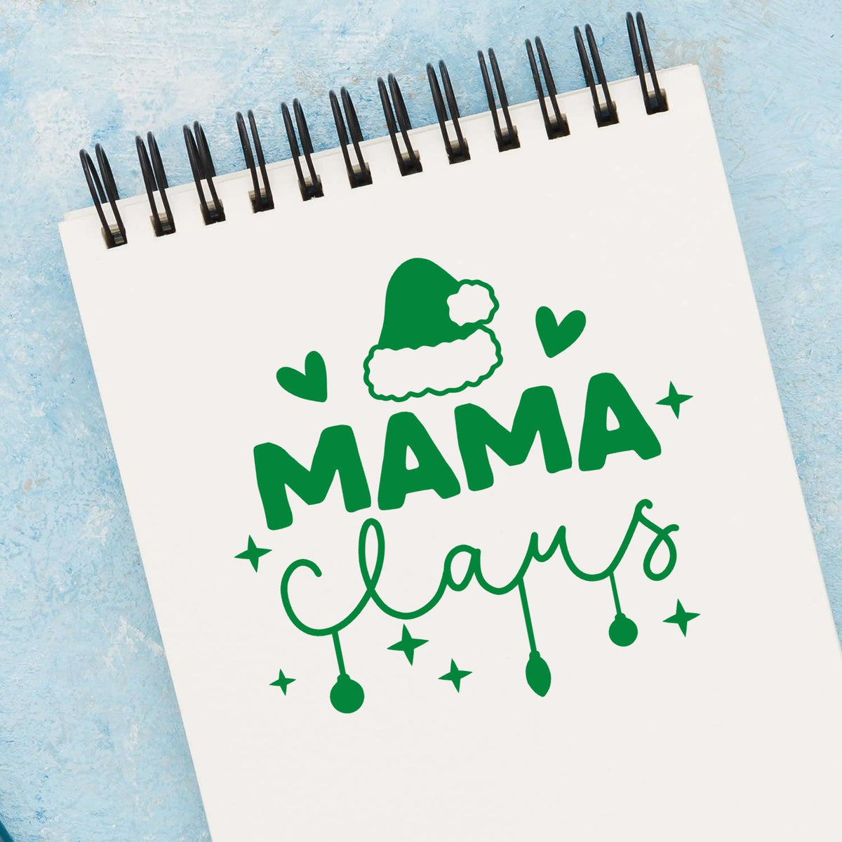 Green MAMA claws stamp and seal design with Santa hat, hearts and stars on a white spiral notebook over blue background.