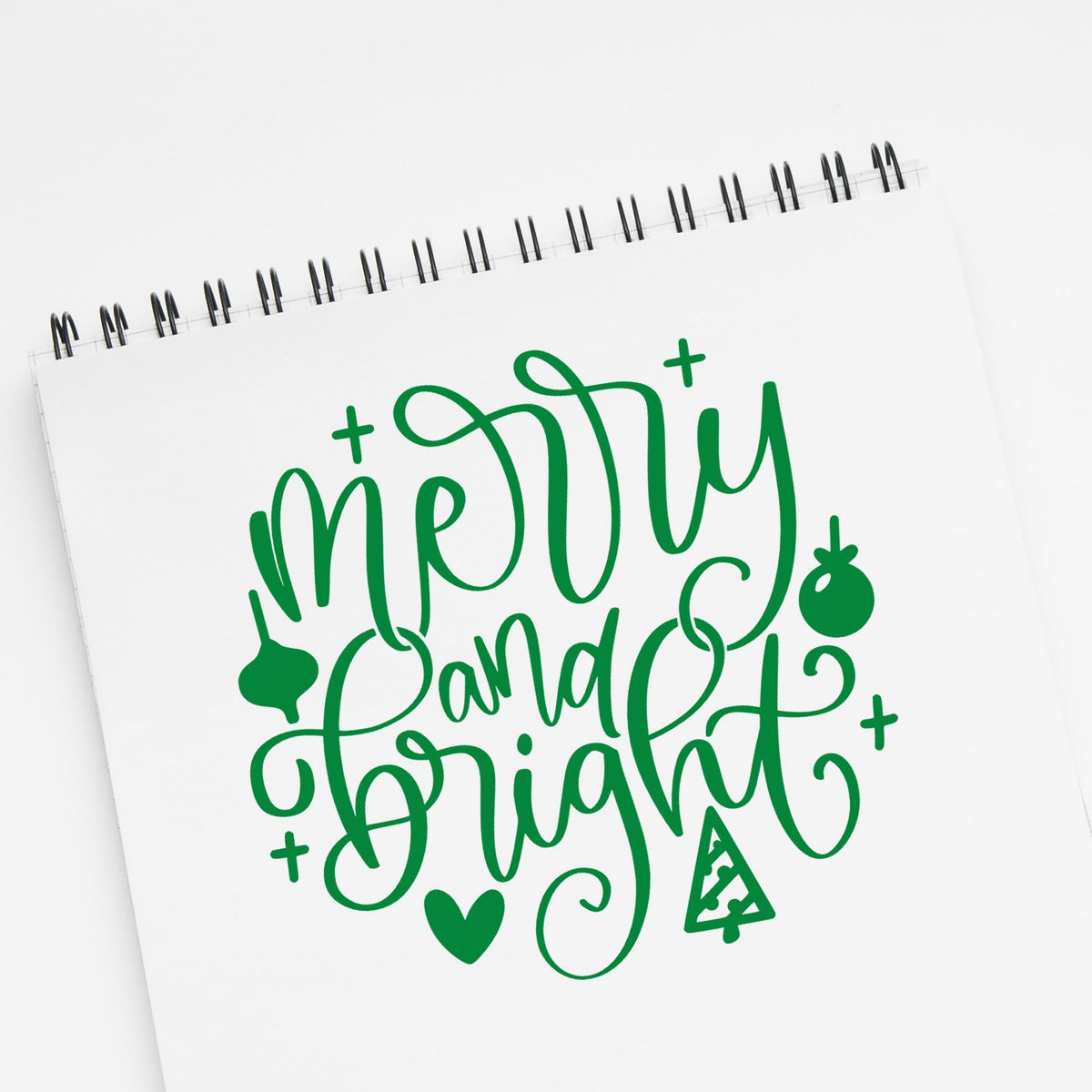 Green Merry and Bright stamp and seal design on white spiral notebook with hearts, tree, and ornament accents.