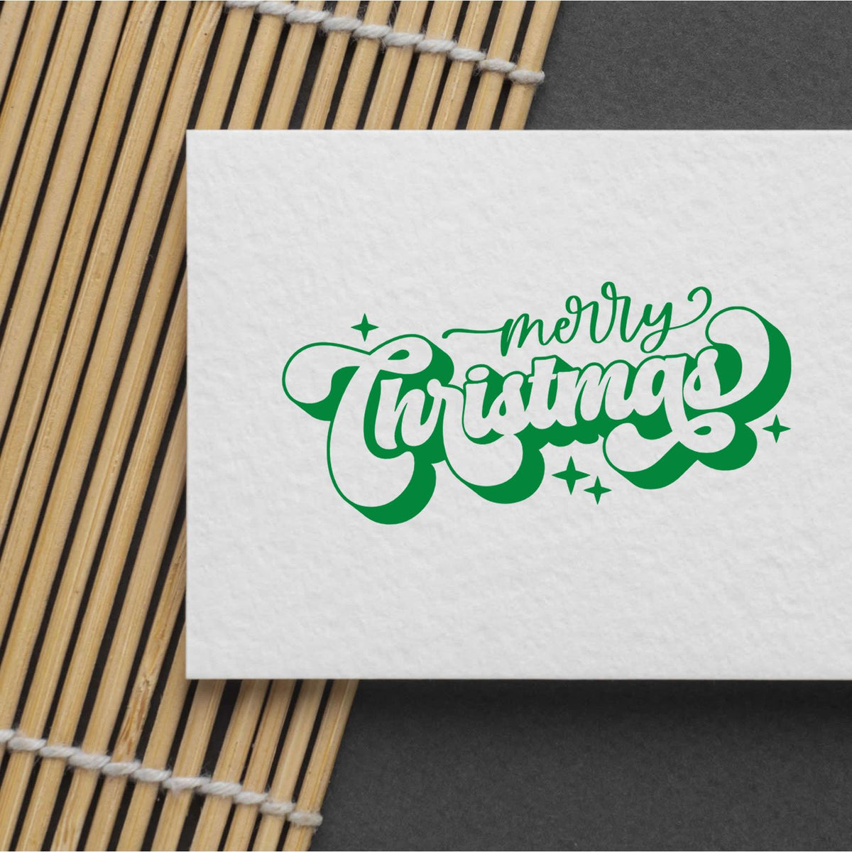 Green Merry Christmas stamp seal on white paper, with bamboo mat and dark gray background.