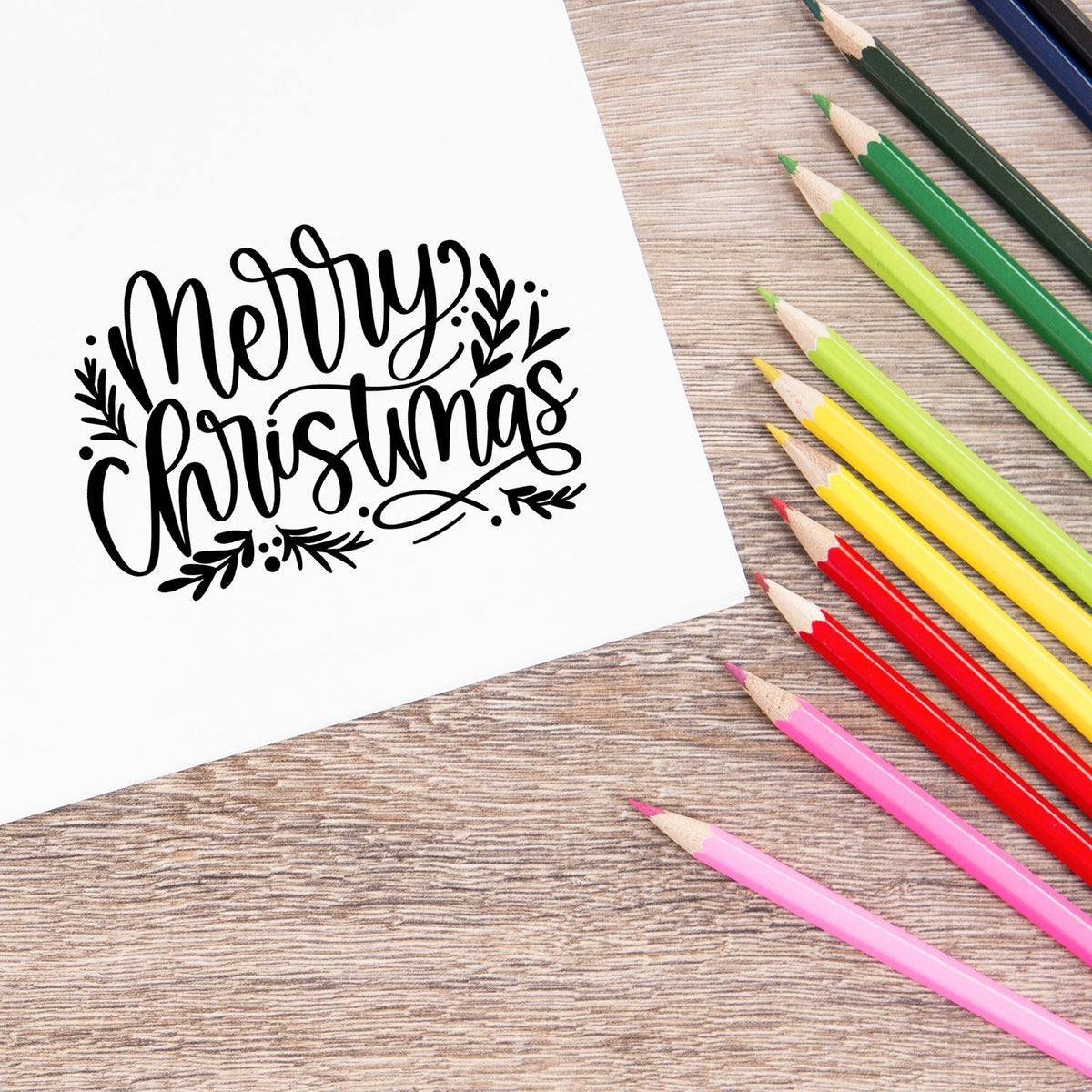 Black Merry Christmas stamp seal on white card, surrounded by colorful pencils on a wood table.