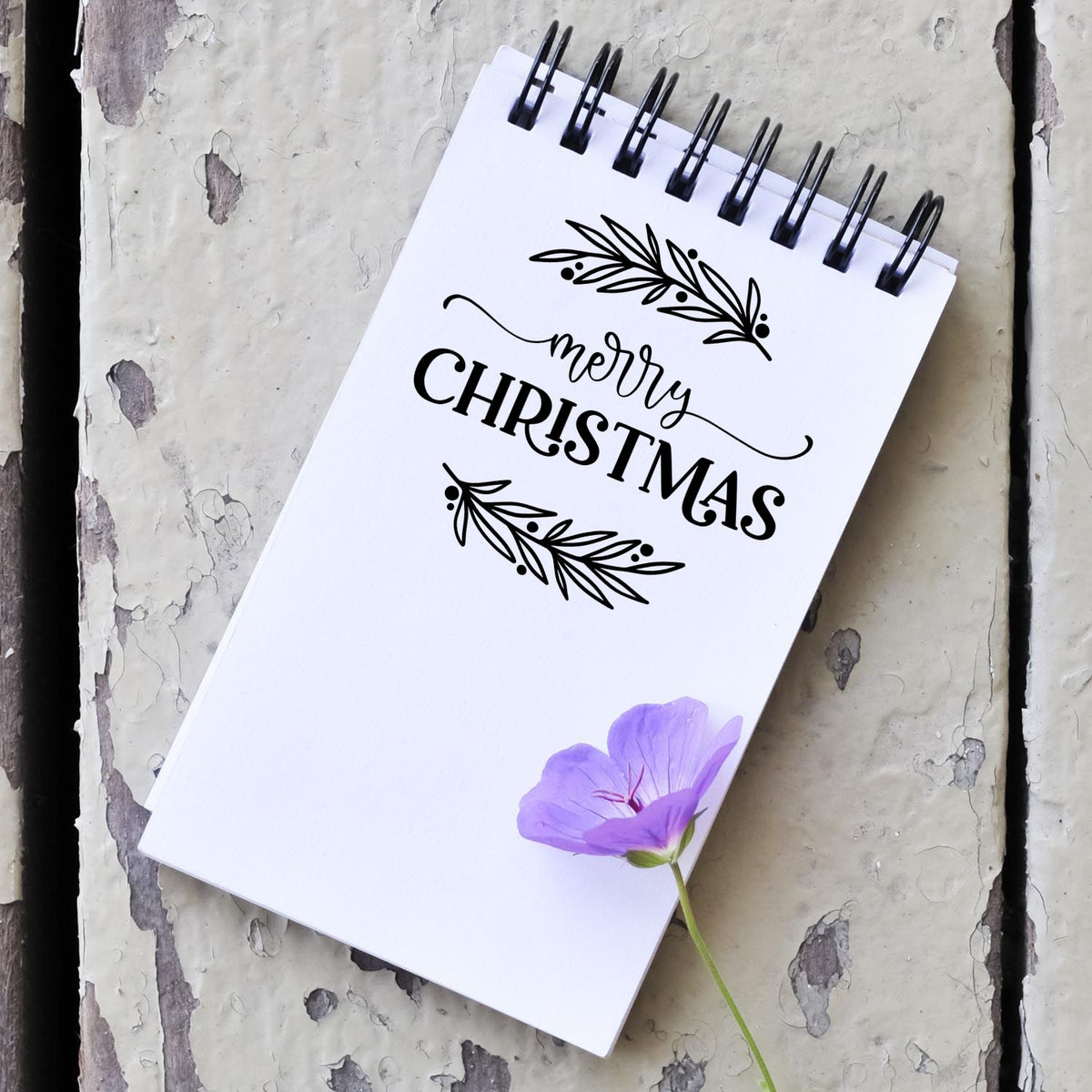 Black Merry Christmas stamp/seal design on white spiral notebook, with olive branches and a purple flower on rustic gray wood.