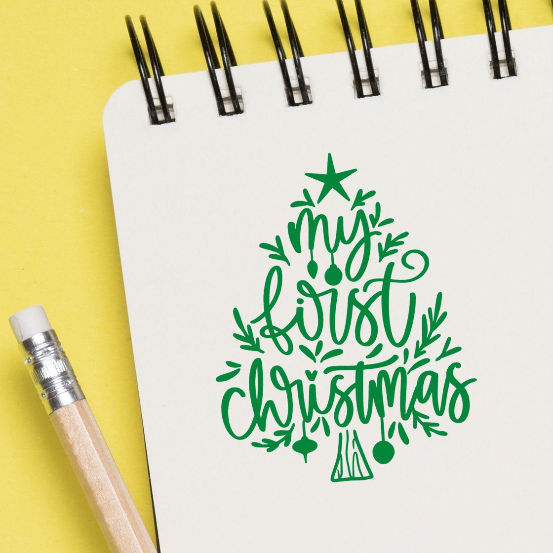 Green Christmas stamp and seal design on white spiral notebook, with yellow background and pencil, reading my first Christmas.