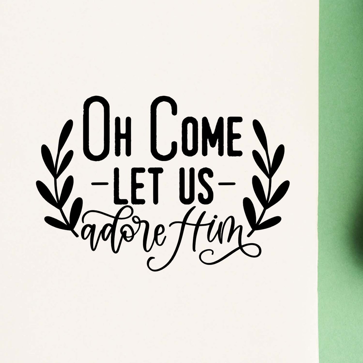 Black holiday stamp seal reading Oh Come Let Us Adore Him, with leafy wreath on white and green background.