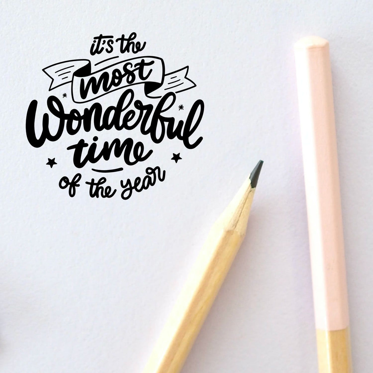 Black custom stamp and seal design reading its the most wonderful time of the year on white paper, with pastel pink and wood pencils.
