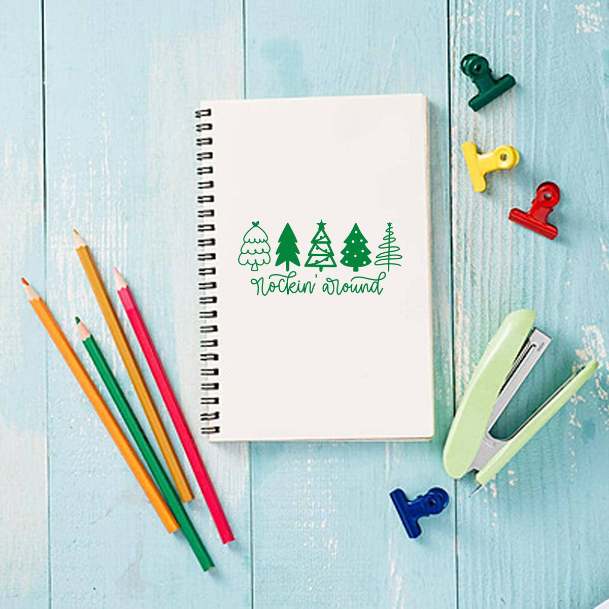 Green Christmas tree stamp and seal on white spiral notebook, with colorful pencils, clips and stapler on blue wood.