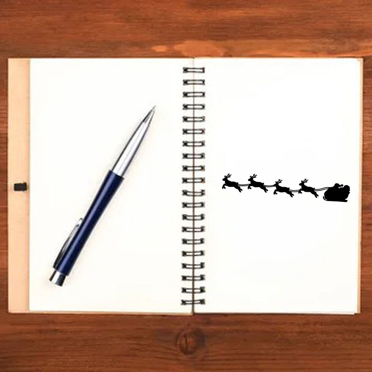 Blue pen on open spiral notebook with black reindeer sleigh stamp and seal on white pages, on a wooden desk.