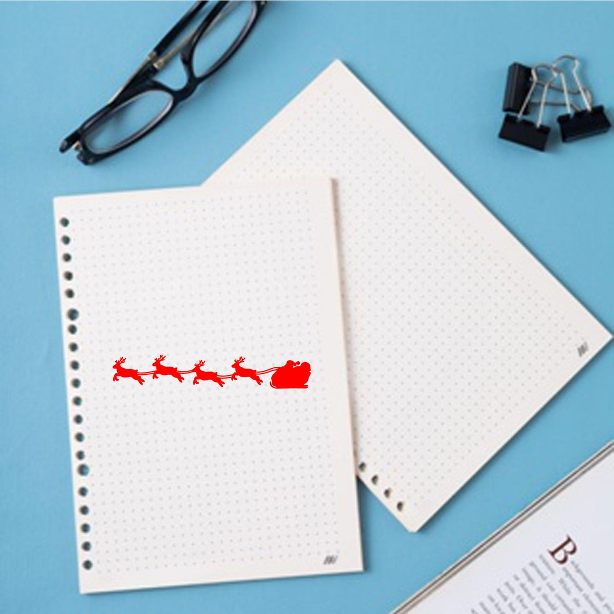Red Santa sleigh stamp and seal on white dotted notebook pages, with black glasses and binder clips on a blue background.