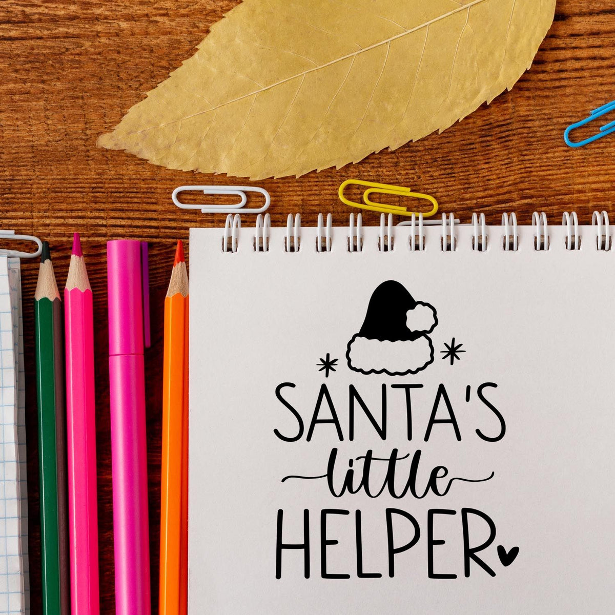 Santas little helper stamp and seal on white spiral notebook, with colorful pencils, clips, and gold leaf on wood desk