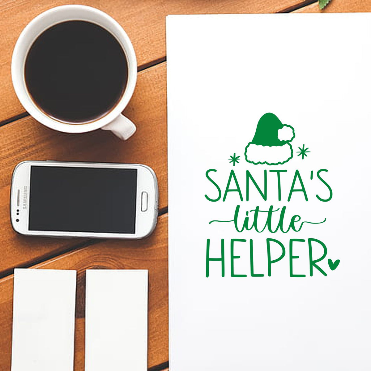 Green Santas Little Helper stamp and seal design on white paper beside coffee, smartphone, and blank cards on wood