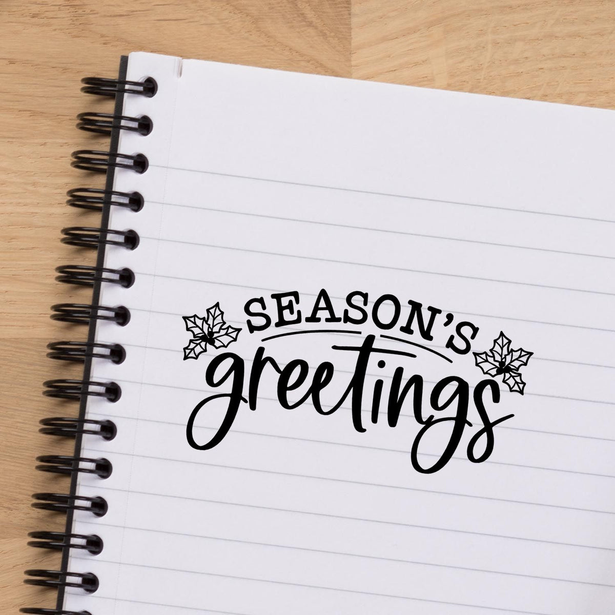 Black holiday stamp and seal reading Seasons greetings with holly on a white lined notebook, black spiral binding on wood.