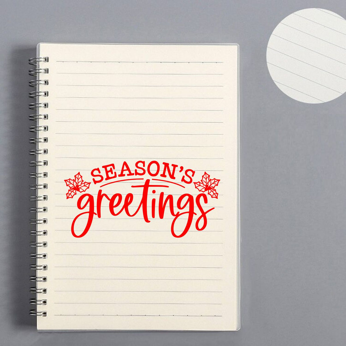 Red Seasons Greetings stamp with holly on cream lined notebook, beside a matching white circular seal on gray.