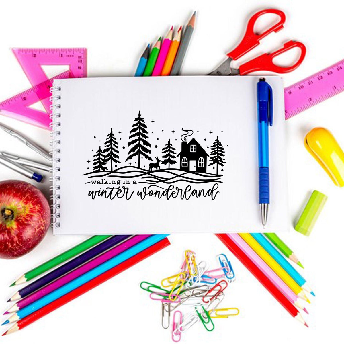 Winter wonderland stamp seal on white notebook, surrounded by colorful pencils, rulers, scissors, pen and paper clips.