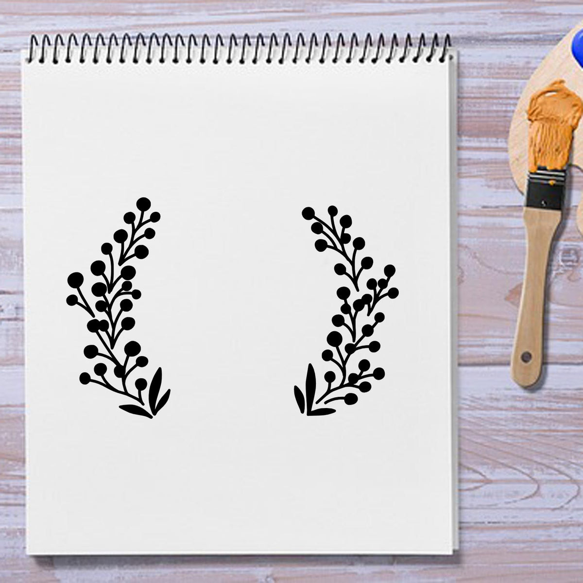 Black botanical stamp seal designs on a white spiral sketchbook, with a paintbrush on a light wood table.
