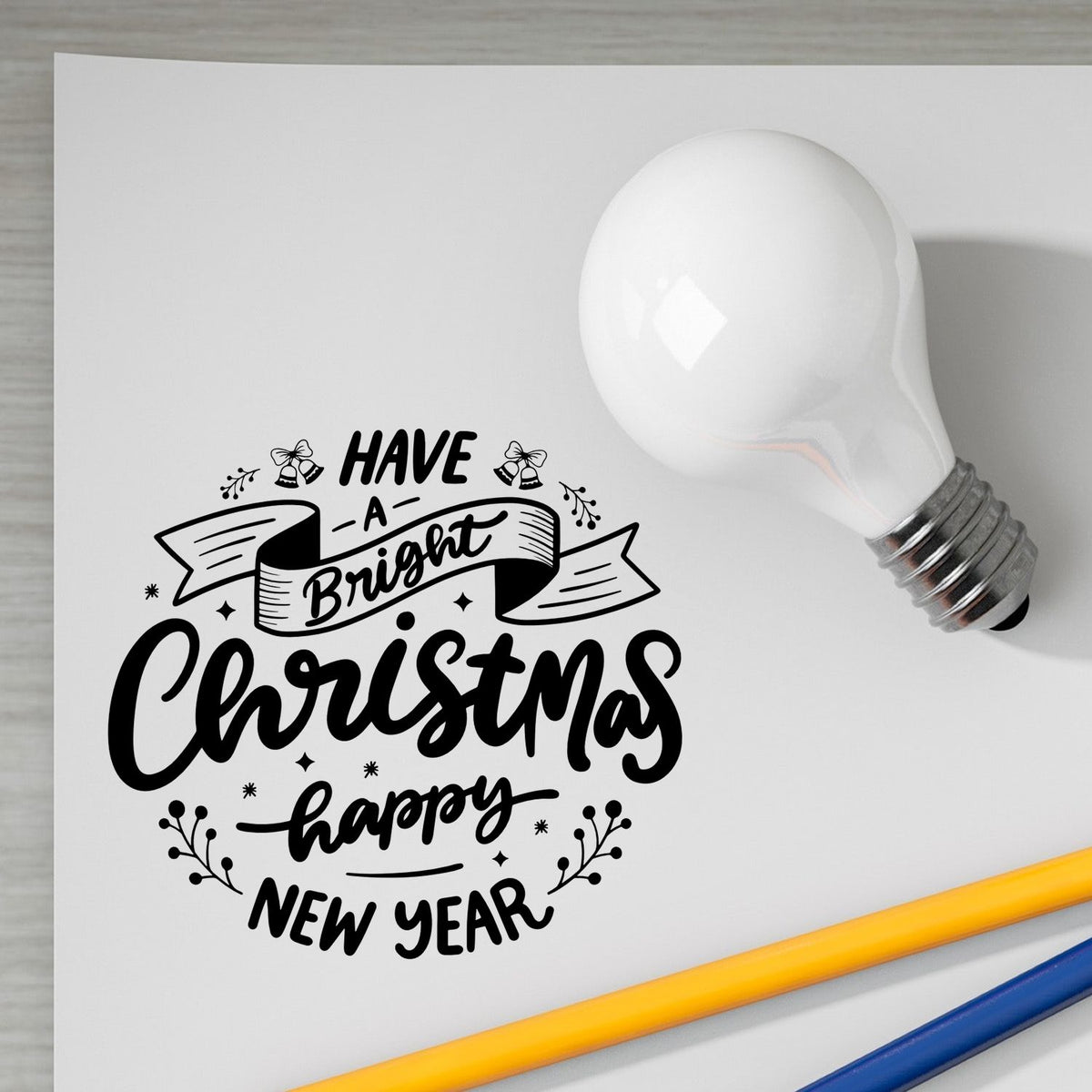 Black Christmas and New Year stamp-and-seal design on white paper beside a white light bulb, with yellow and blue pencils.