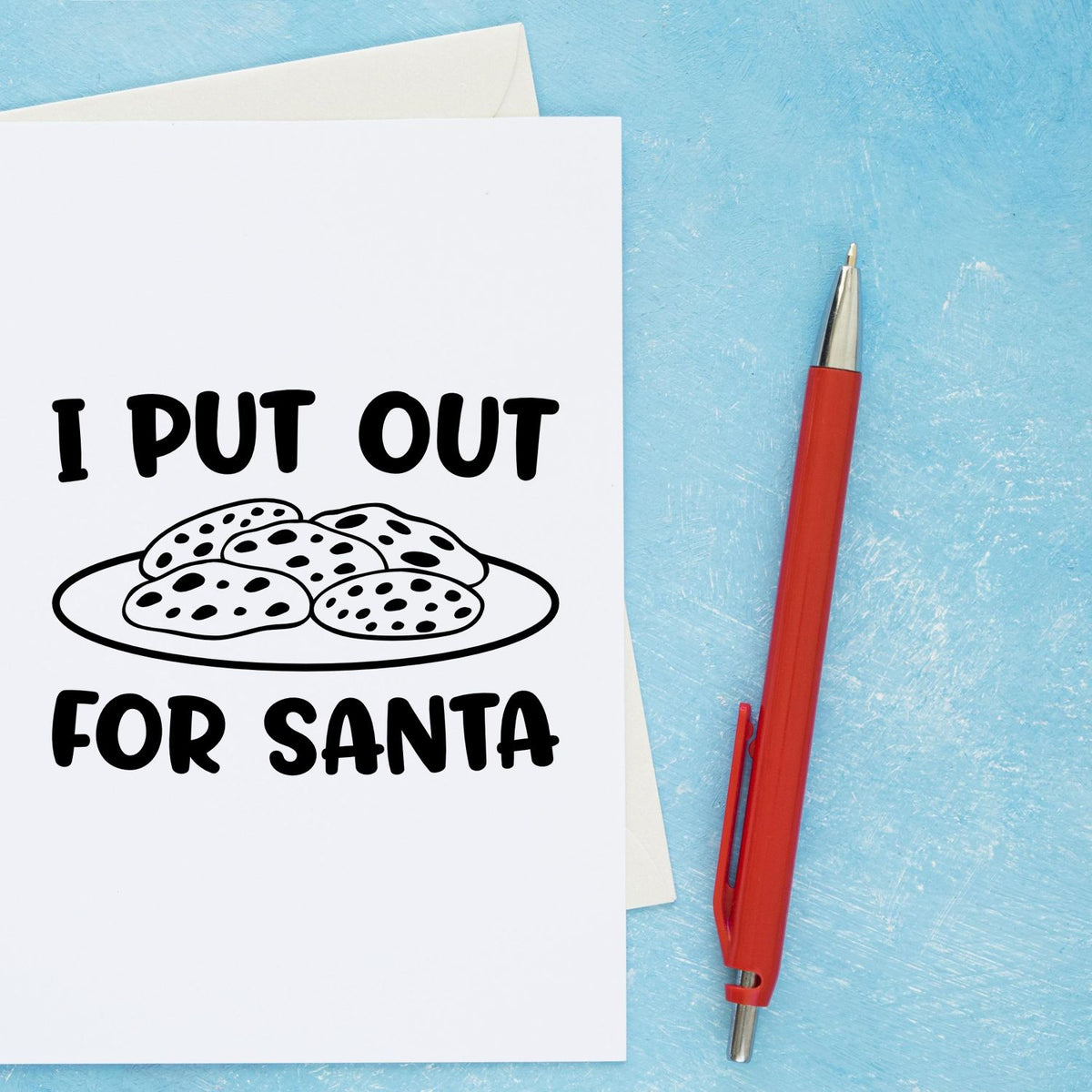 Red self-inking stamp pen beside a white holiday card reading I PUT OUT FOR SANTA with cookie graphic on a blue background.