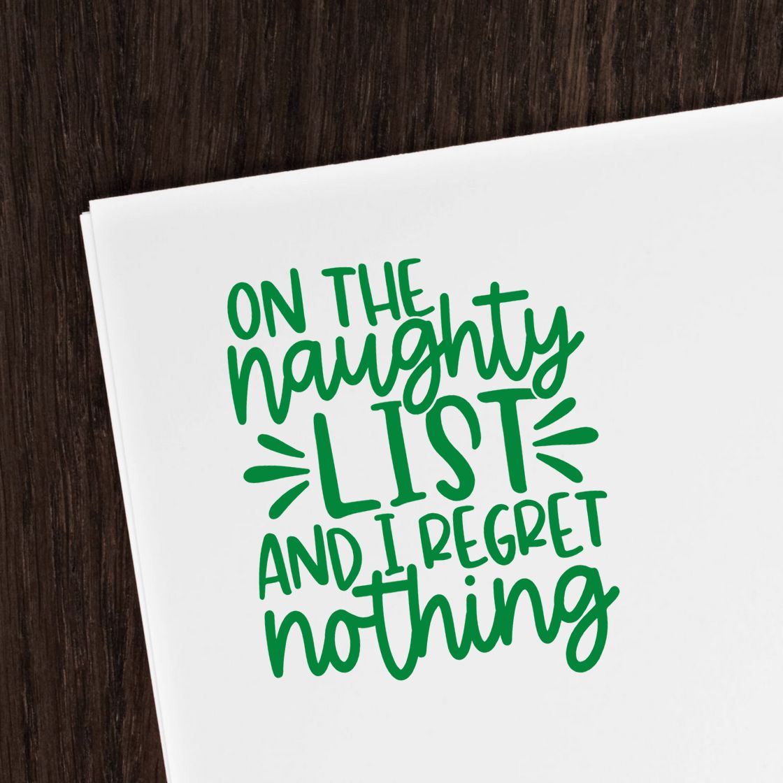 Green stamp and seal design on white paper reading On the naughty list and I regret nothing over dark wood.