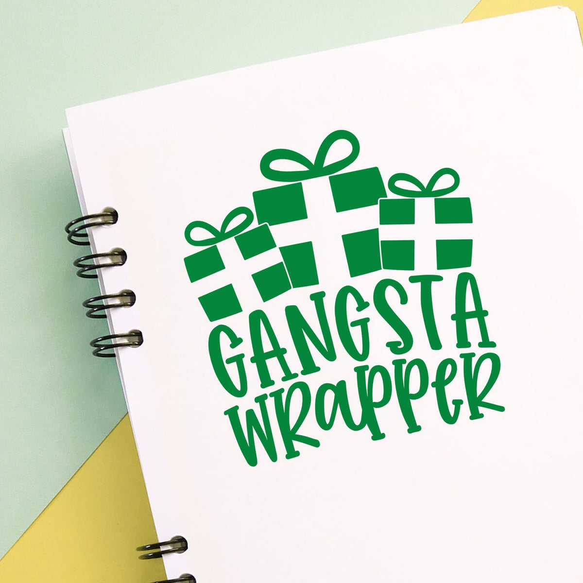 Green gangsta wrapper gift stamp and seal on white spiral notebook with pastel green and yellow background.