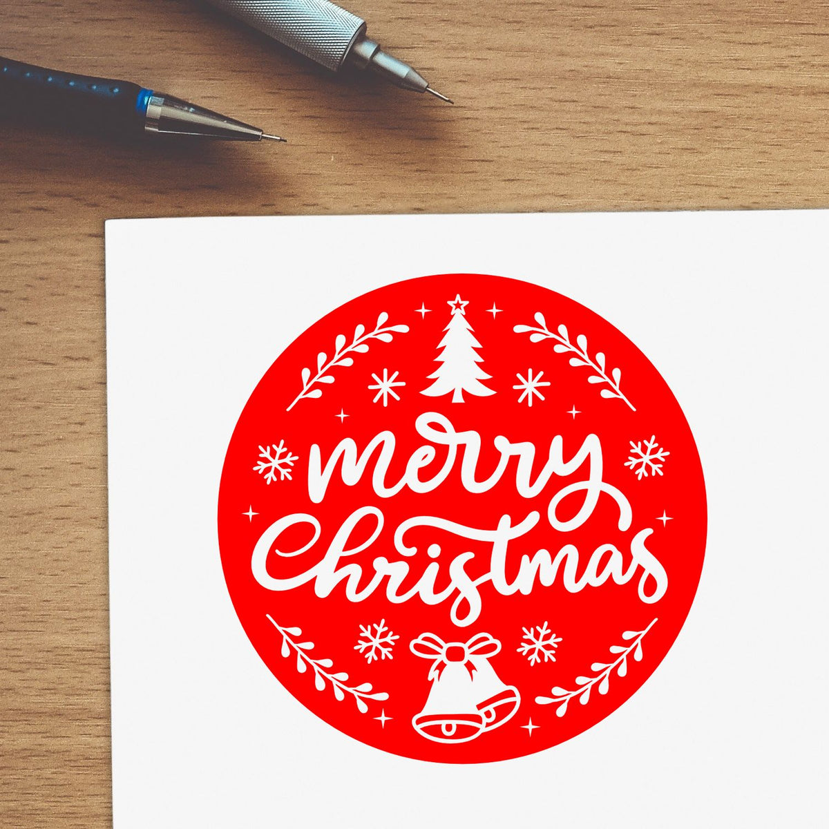 Red Merry Christmas stamp seal with white tree, bells and snowflakes on white paper beside pens on a wood desk.
