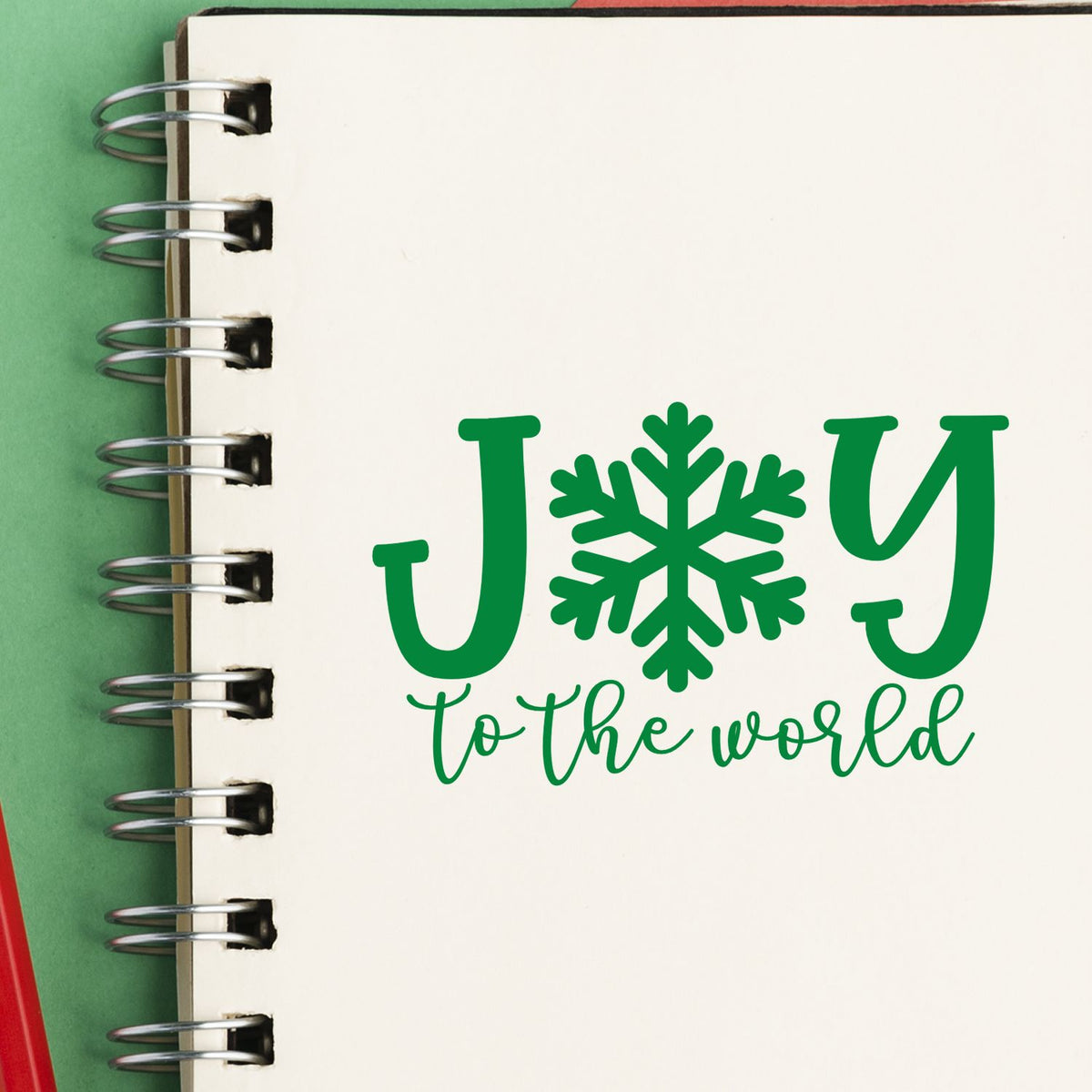 Green holiday stamp and seal design on cream notebook, reading JOY to the world with a snowflake.
