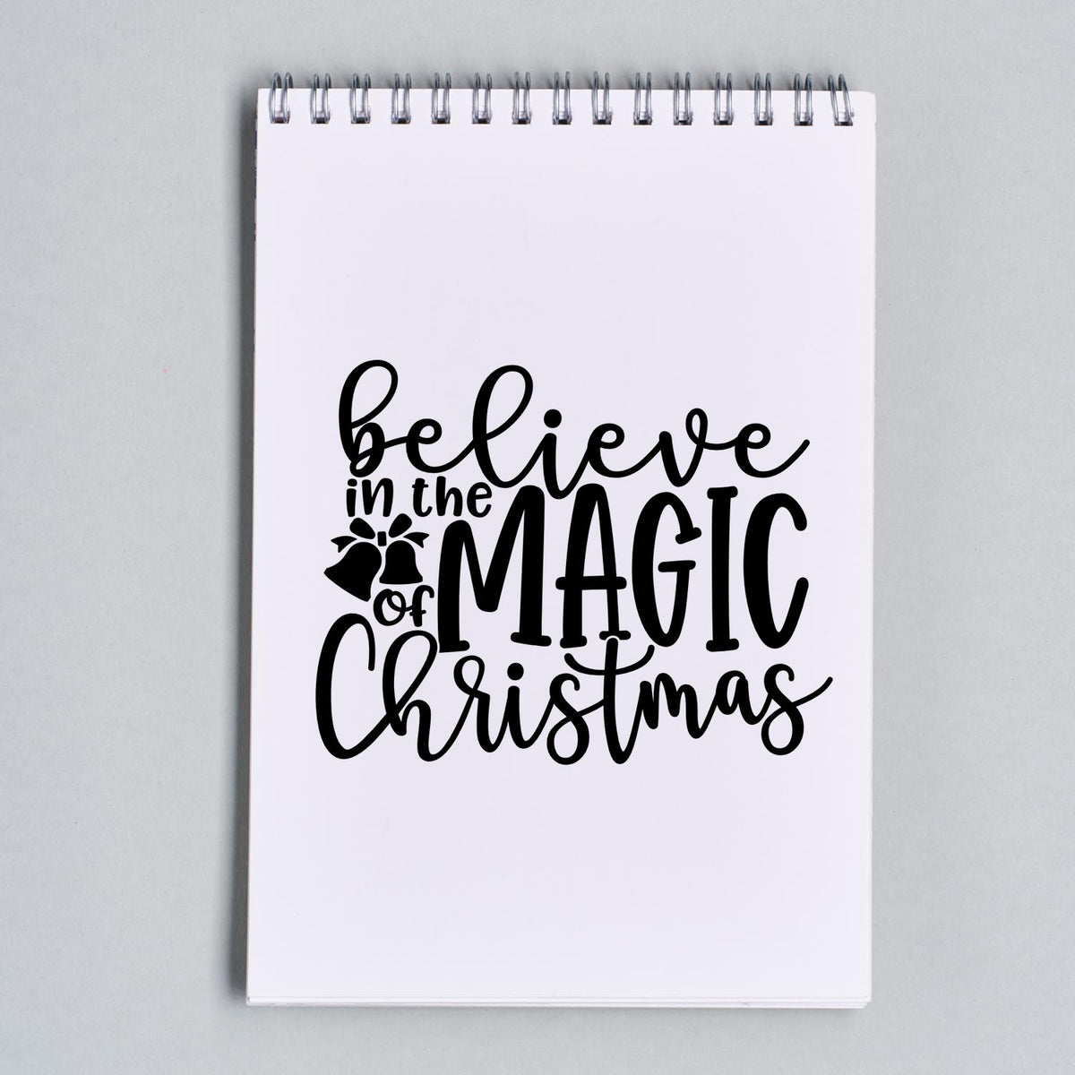 Black Christmas stamp and seal design on a white spiral notebook, reading Believe in the Magic of Christmas with bells.