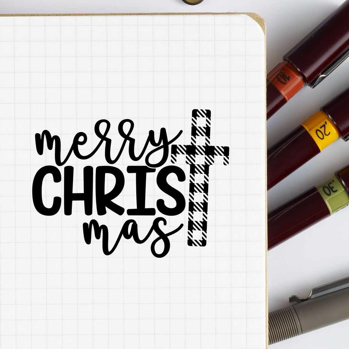 Black Merry Christmas lettering with plaid cross on grid paper, beside burgundy stamp and seal tools with colored bands.