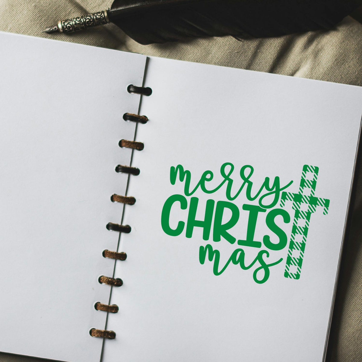 Green Merry Christmas stamp and plaid seal on white spiral notebook, with black feather and pen