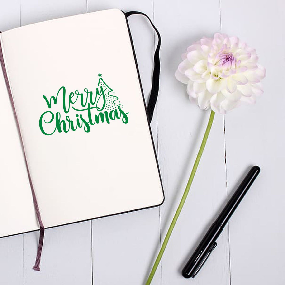 White notebook with green Merry Christmas stamp and seal, black pen, and pale pink flower on a white wood background
