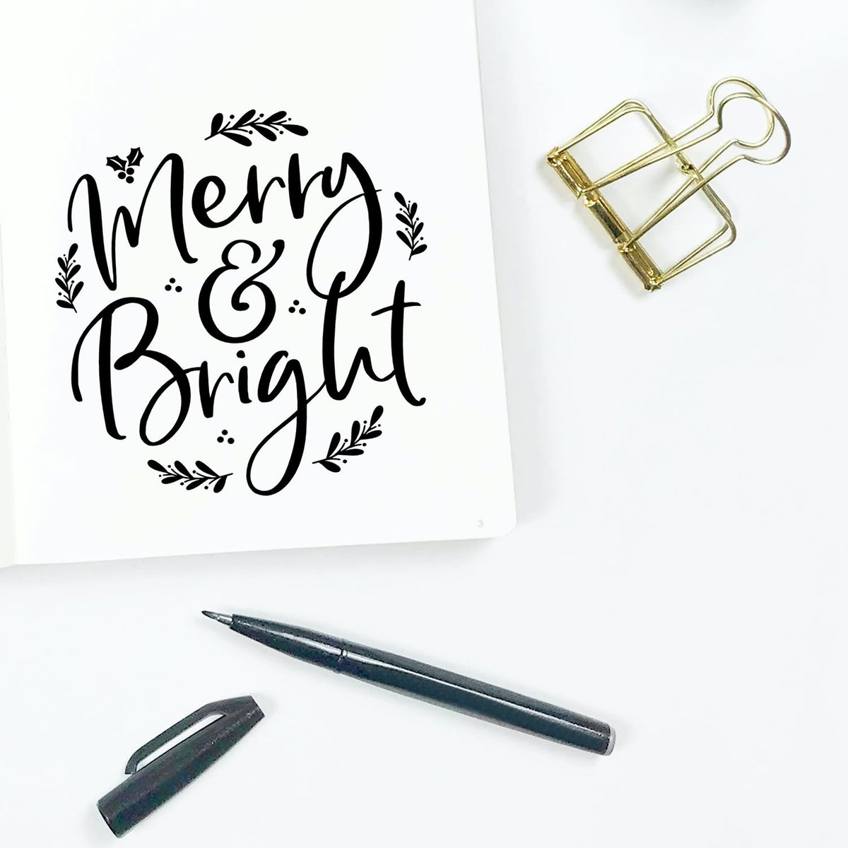 Black Merry & Bright stamp and seal design on white paper, with gold binder clip, black pen and cap.
