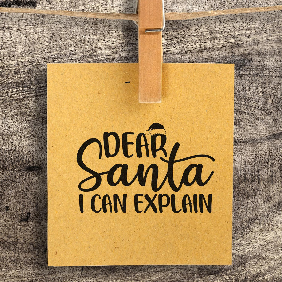 Yellow hanging stamp seal with black Dear Santa I can explain text, Santa hat detail, clipped to rustic wood background.