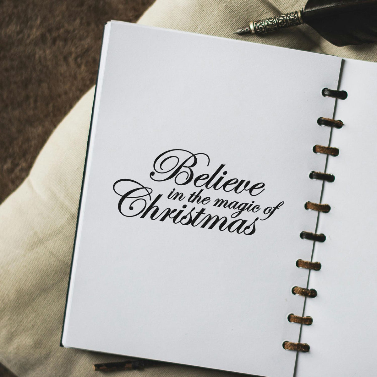 Custom black stamp and seal on white notebook reads Believe in the magic of Christmas, with vintage pen and leather binding.