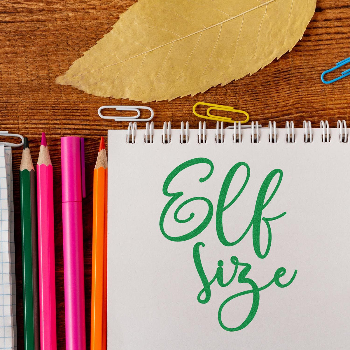 Green Elf size stamp and seal design on a white spiral notebook, with pink, orange, and green pencils on a wood desk.