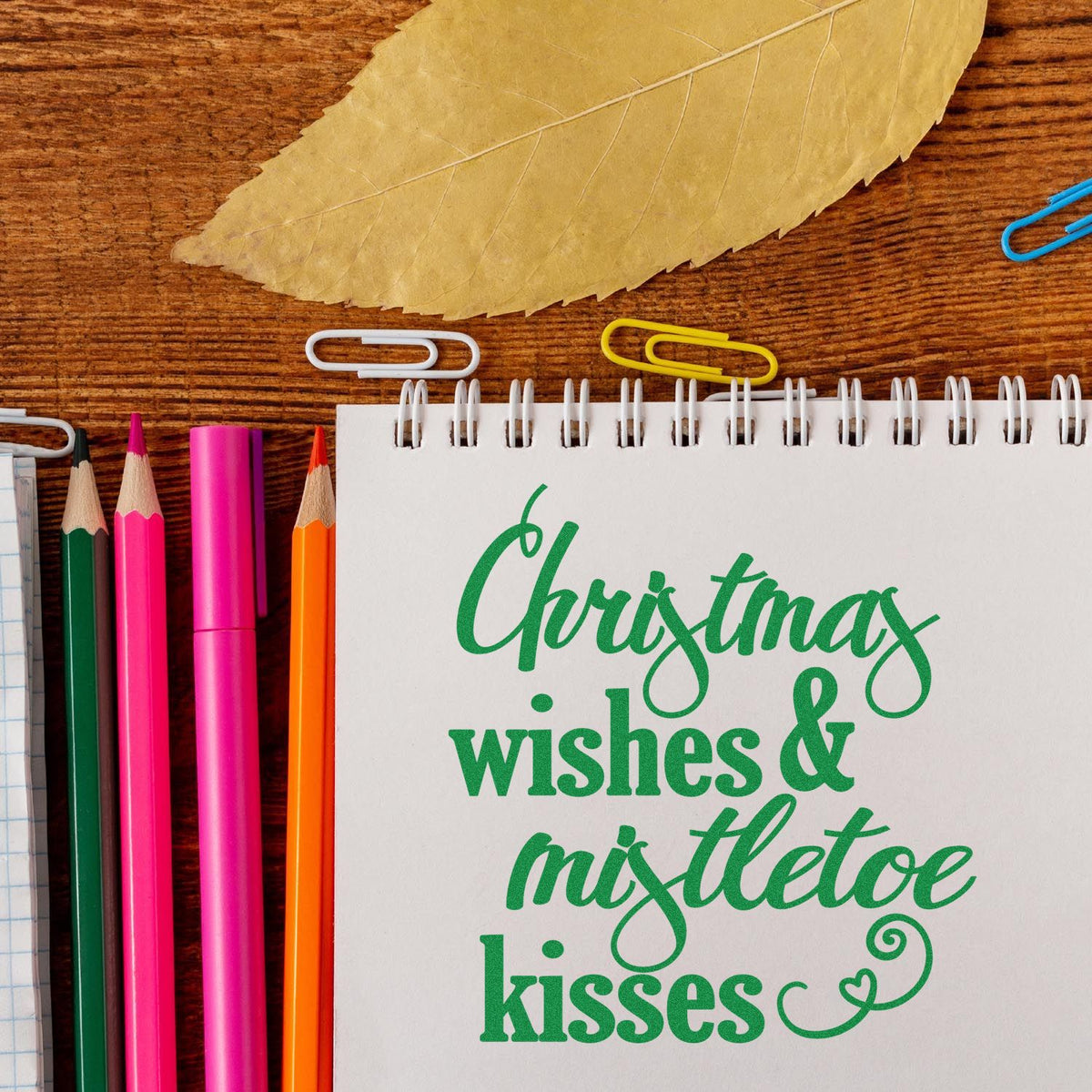Green Christmas wishes stamp and seal on a white spiral notebook, with colorful pencils, paper clips, and a leaf on wood.