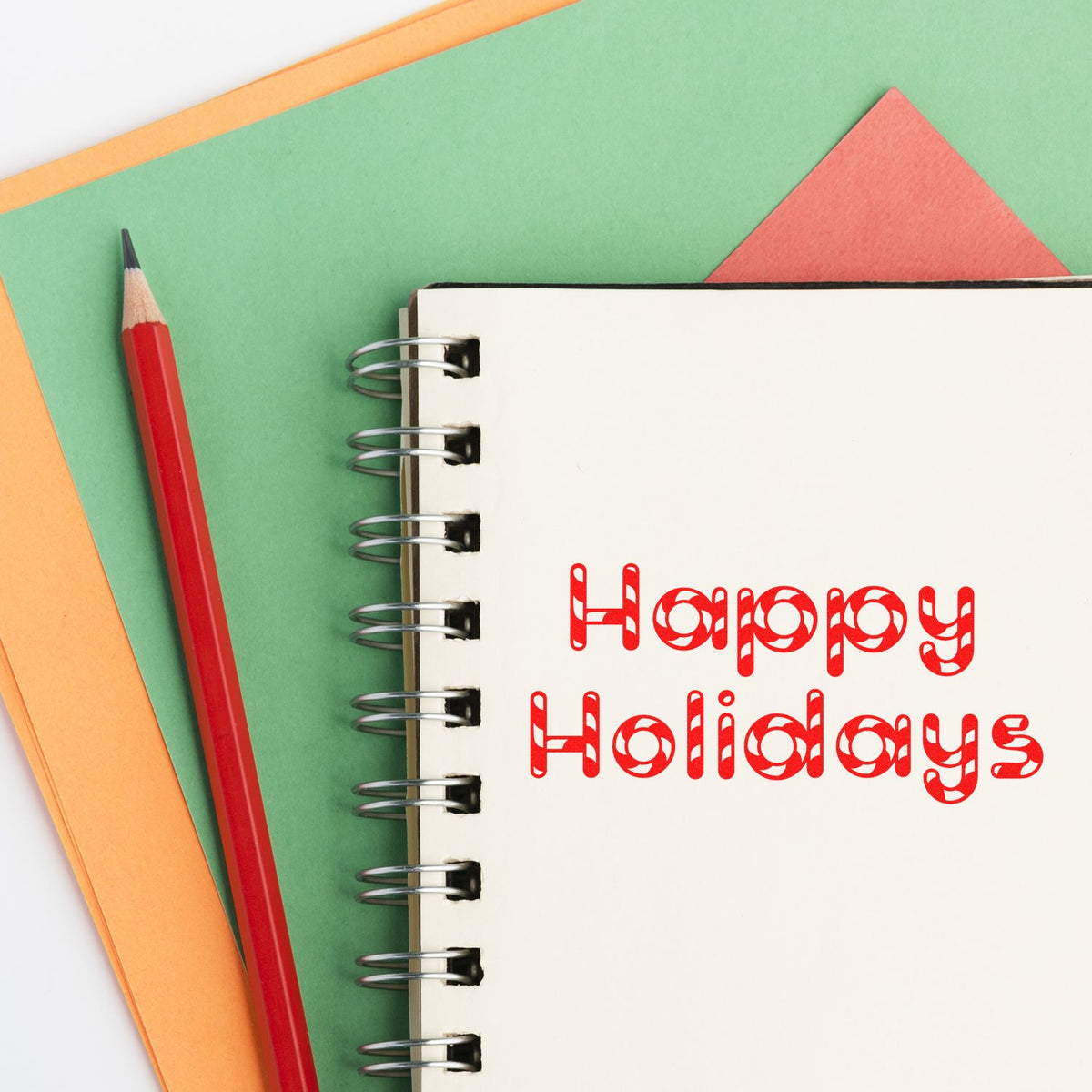 Red Happy Holidays stamp-and-seal design on a spiral notebook, with a red pencil on green and orange paper.