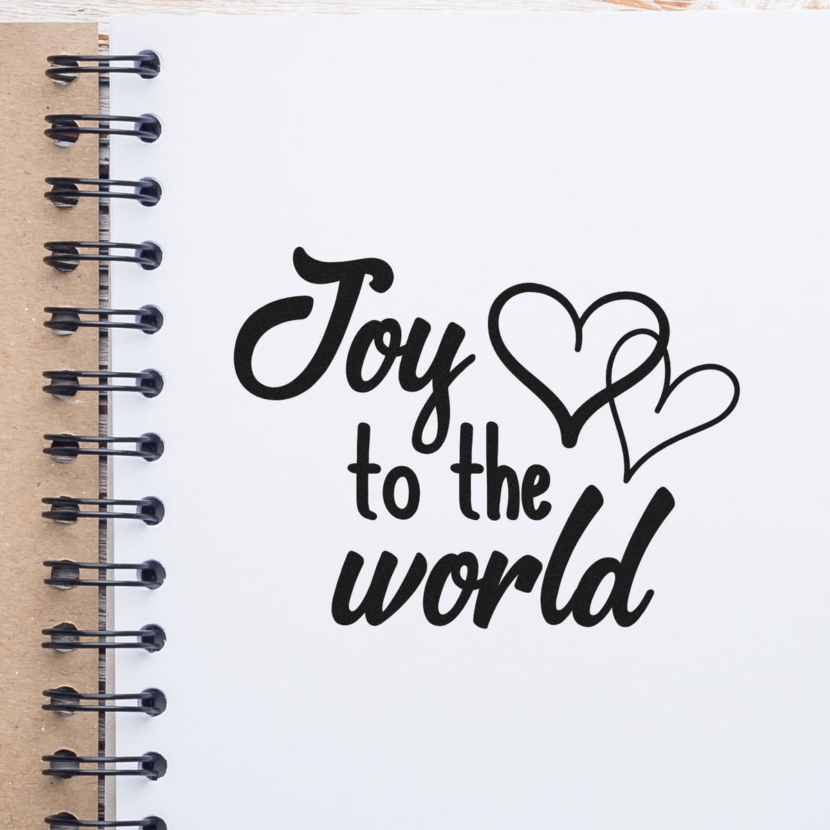 Black Joy to the world stamp design with heart accents on a white spiral notebook, brown kraft spine, and black wire binding.