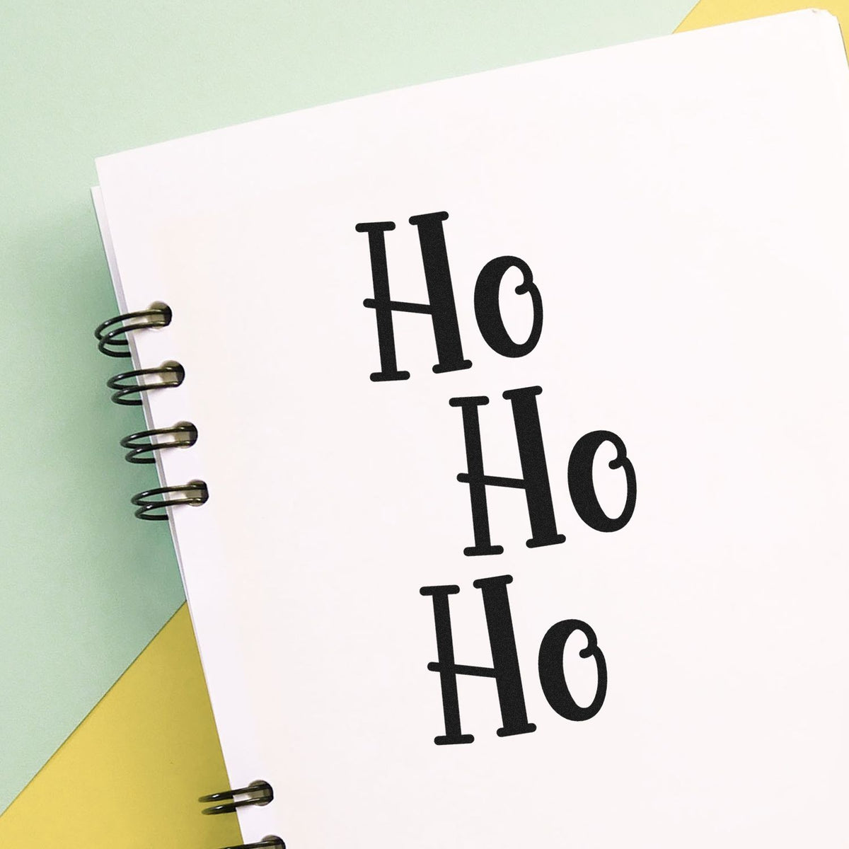 Black Ho Ho Ho stamp seal on white spiral-bound notebook pages, with mint green and yellow background.