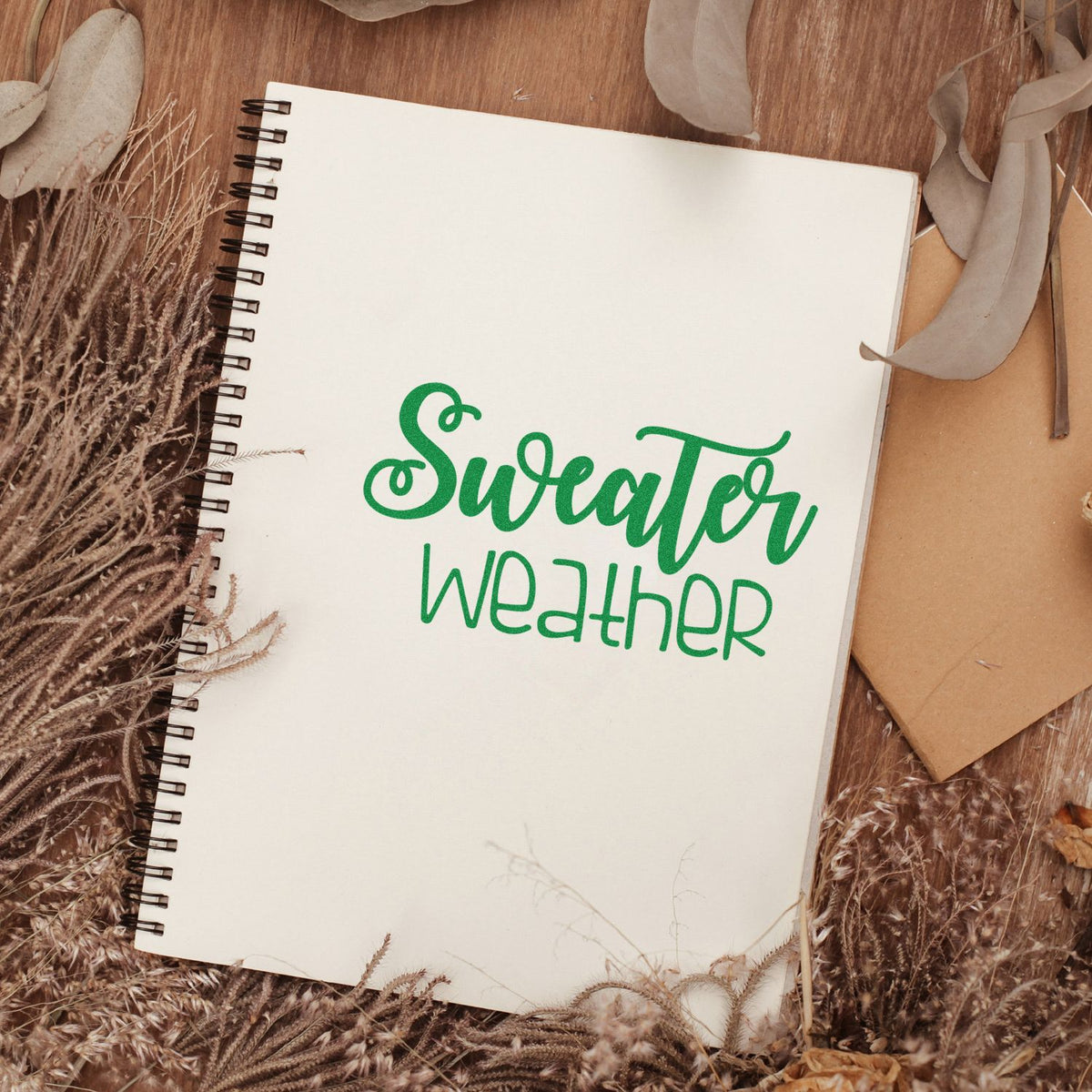 Green Sweater Weather stamp/seal on a white spiral notebook, framed by dried grasses and brown kraft paper accents.
