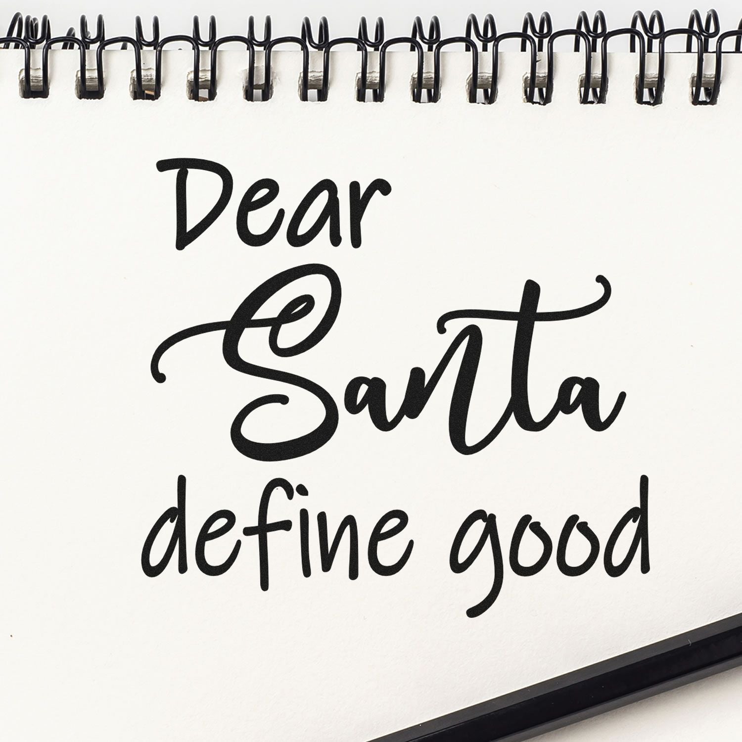 Black script stamp and seal design on white spiral notebook reading Dear Santa define good.