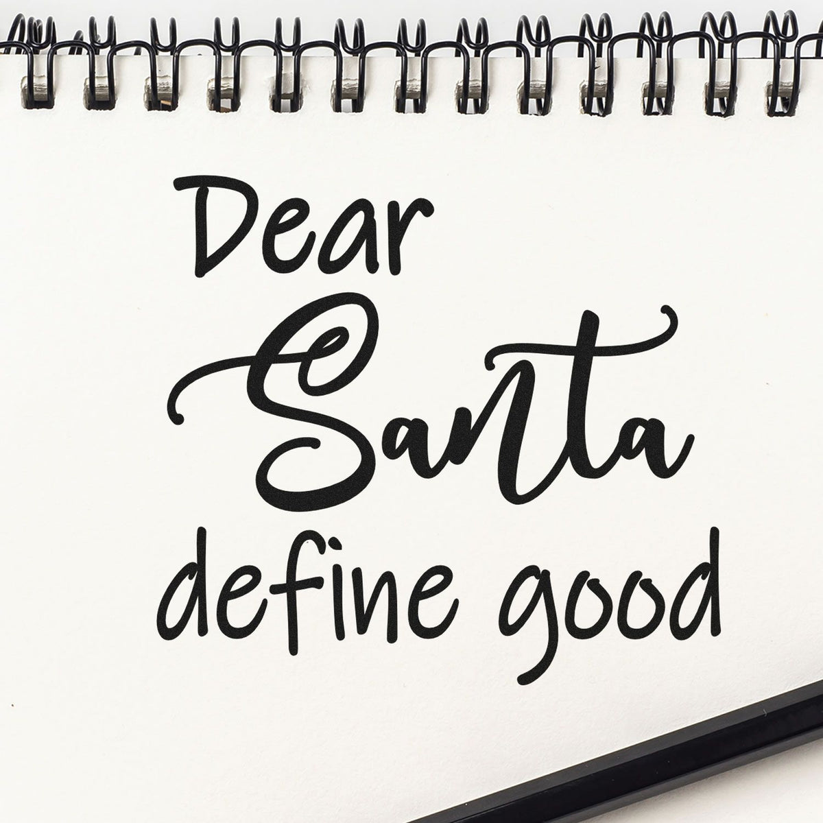 Black stamp and seal design on a white spiral notebook reading Dear Santa define good in bold script.