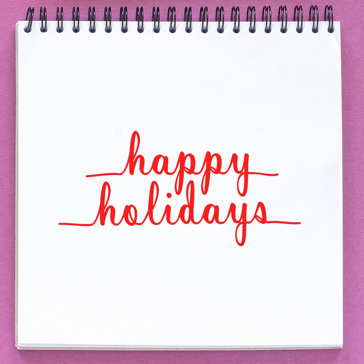 Red Happy Holidays rubber stamp on a white spiral notebook, with black binding and a pink background.