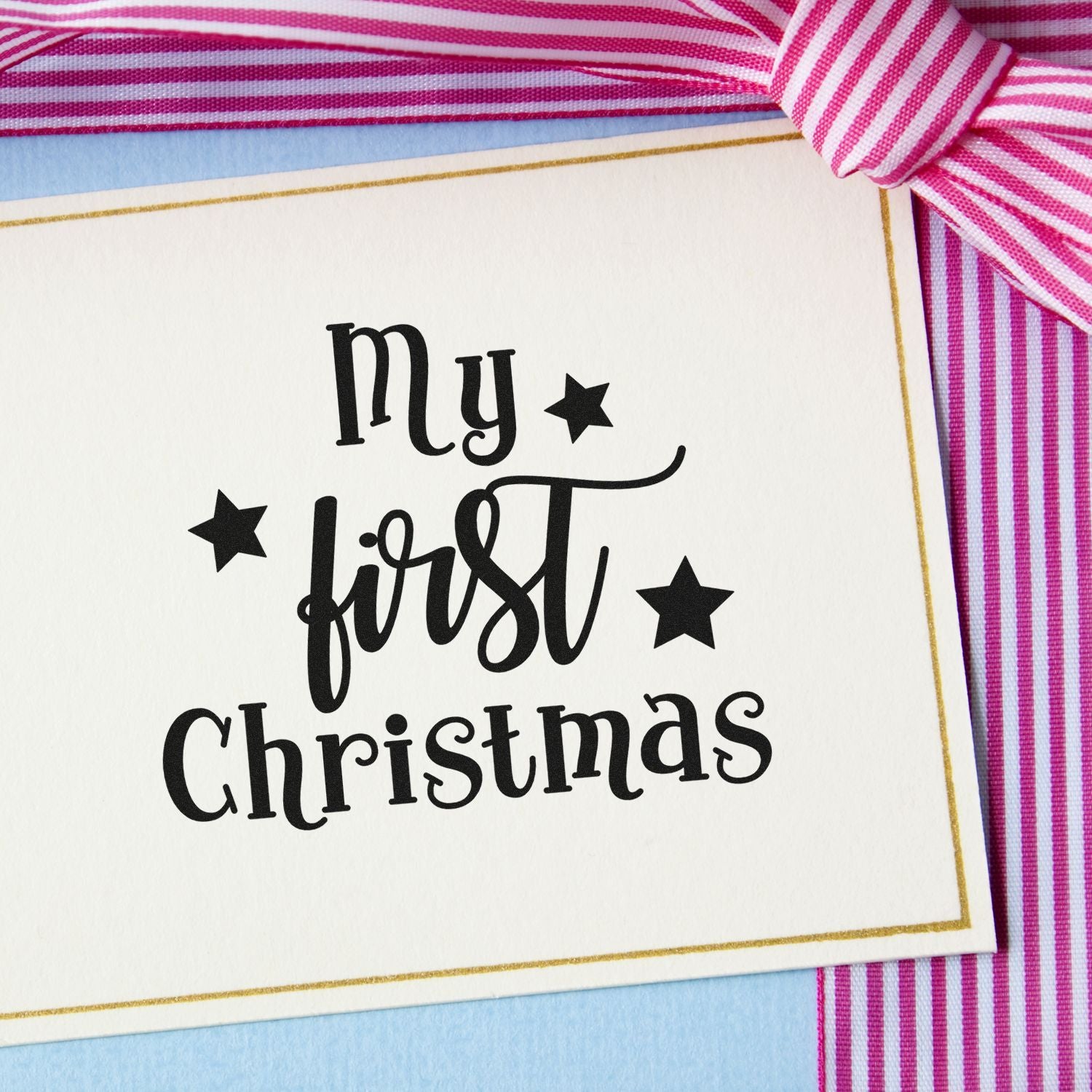 Holiday stamp and seal reading My first Christmas in black on cream card, with gold border, stars, pink ribbon, and blue backing.