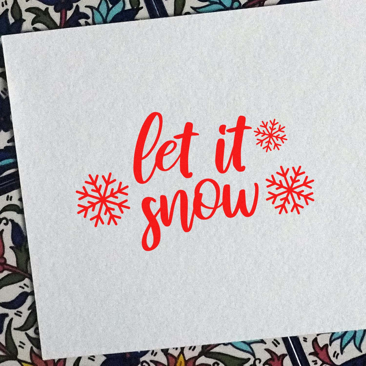 Red holiday stamp and seal design reading let it snow with snowflakes on white paper.