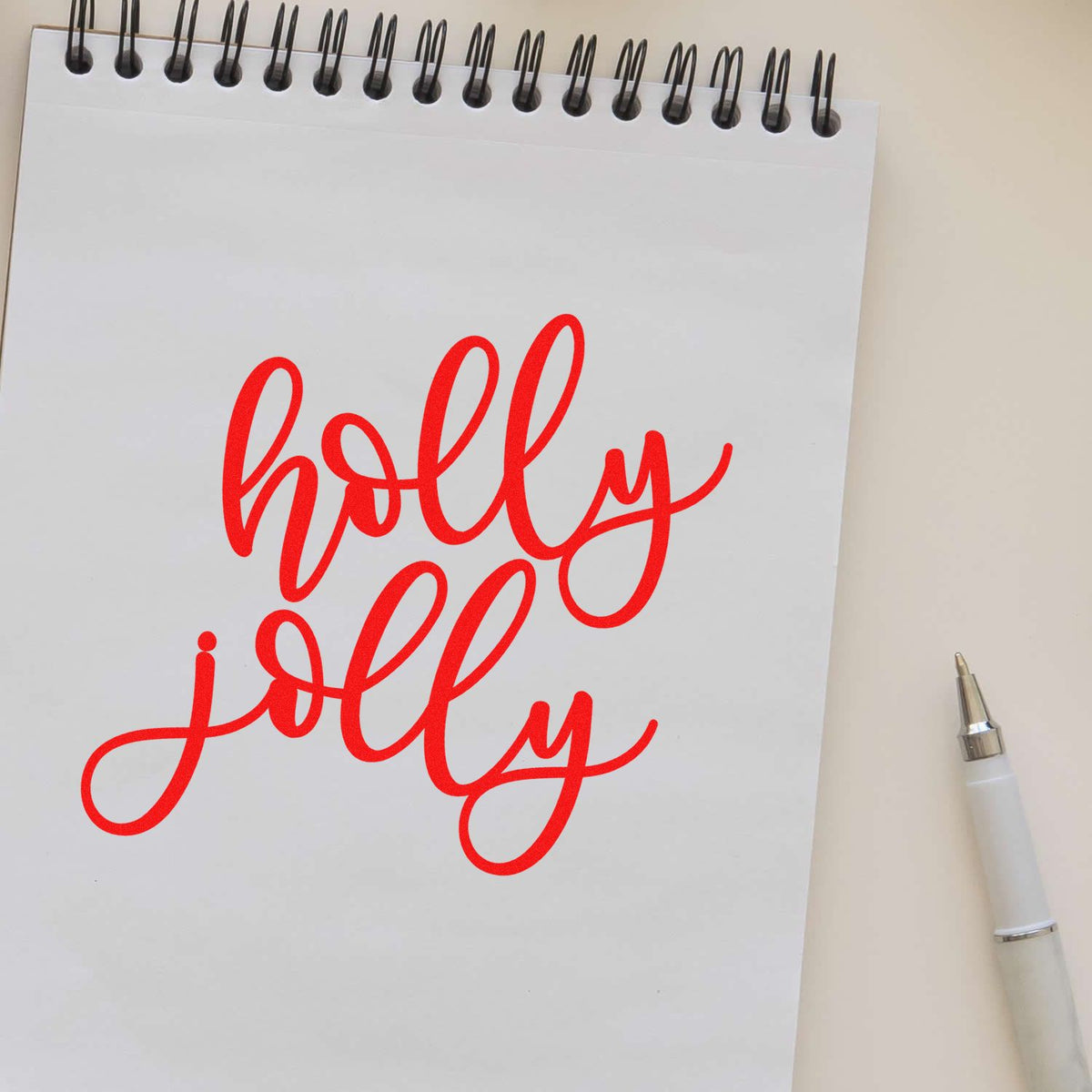 Red holly jolly stamp and seal script on white spiral notepad, with silver pen on beige background.