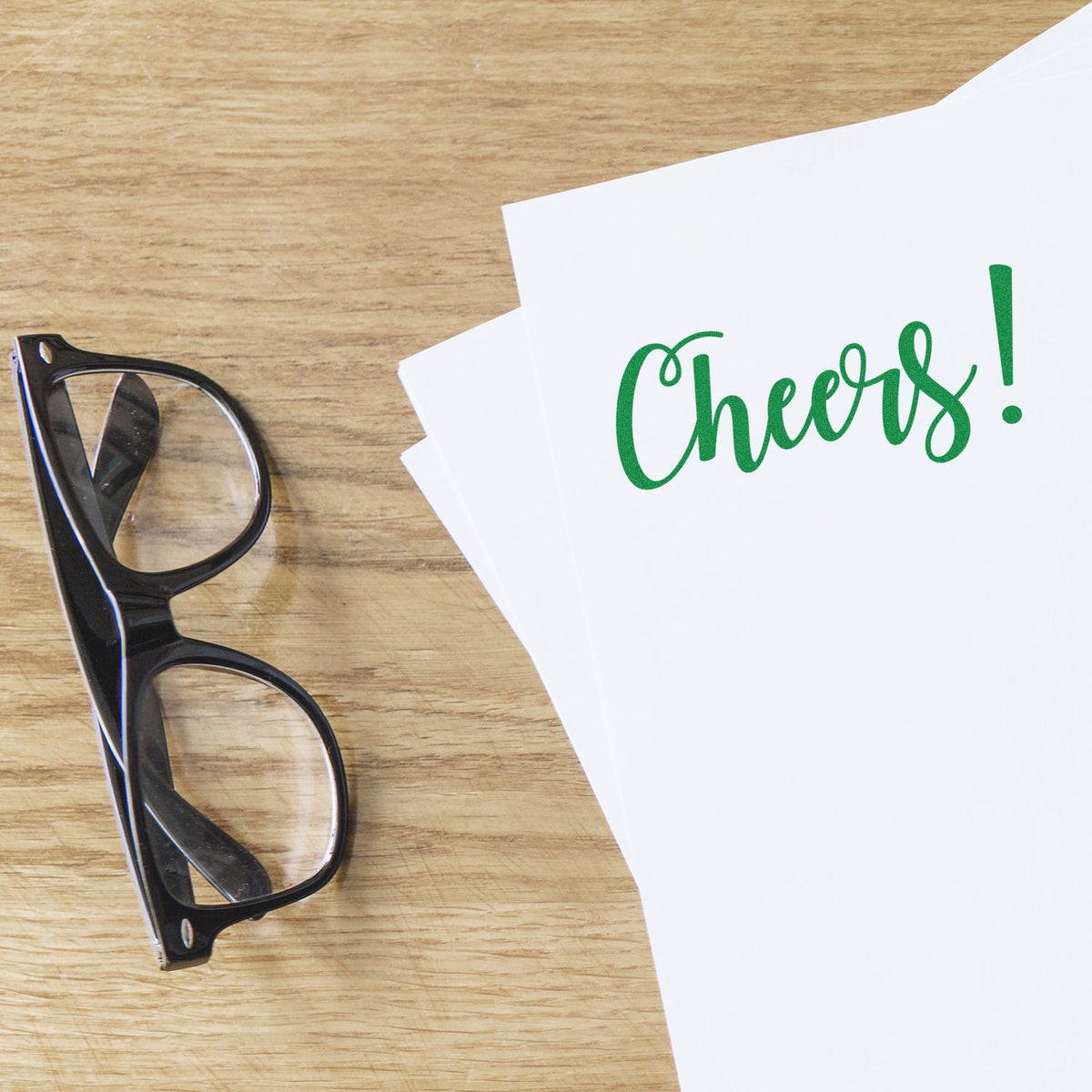 Black glasses beside white paper stamped with green Cheers! on a wood desk, featuring stamp and seal products.