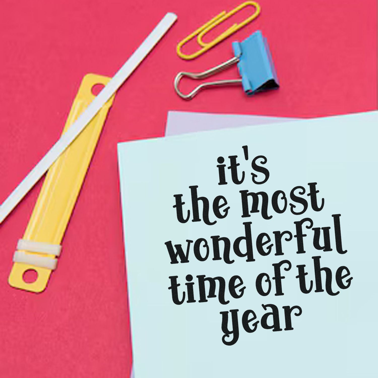 Pink backdrop with stamp and seal supplies, yellow utility knife, blue binder clip, and a card reading its the most wonderful time of the year.