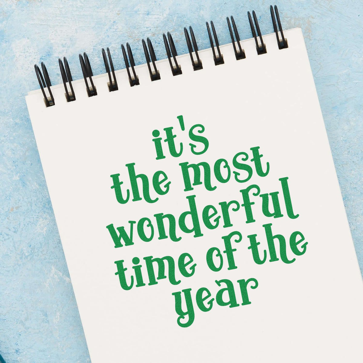 Green holiday stamp on white spiral notebook reads its the most wonderful time of the year on a blue background.