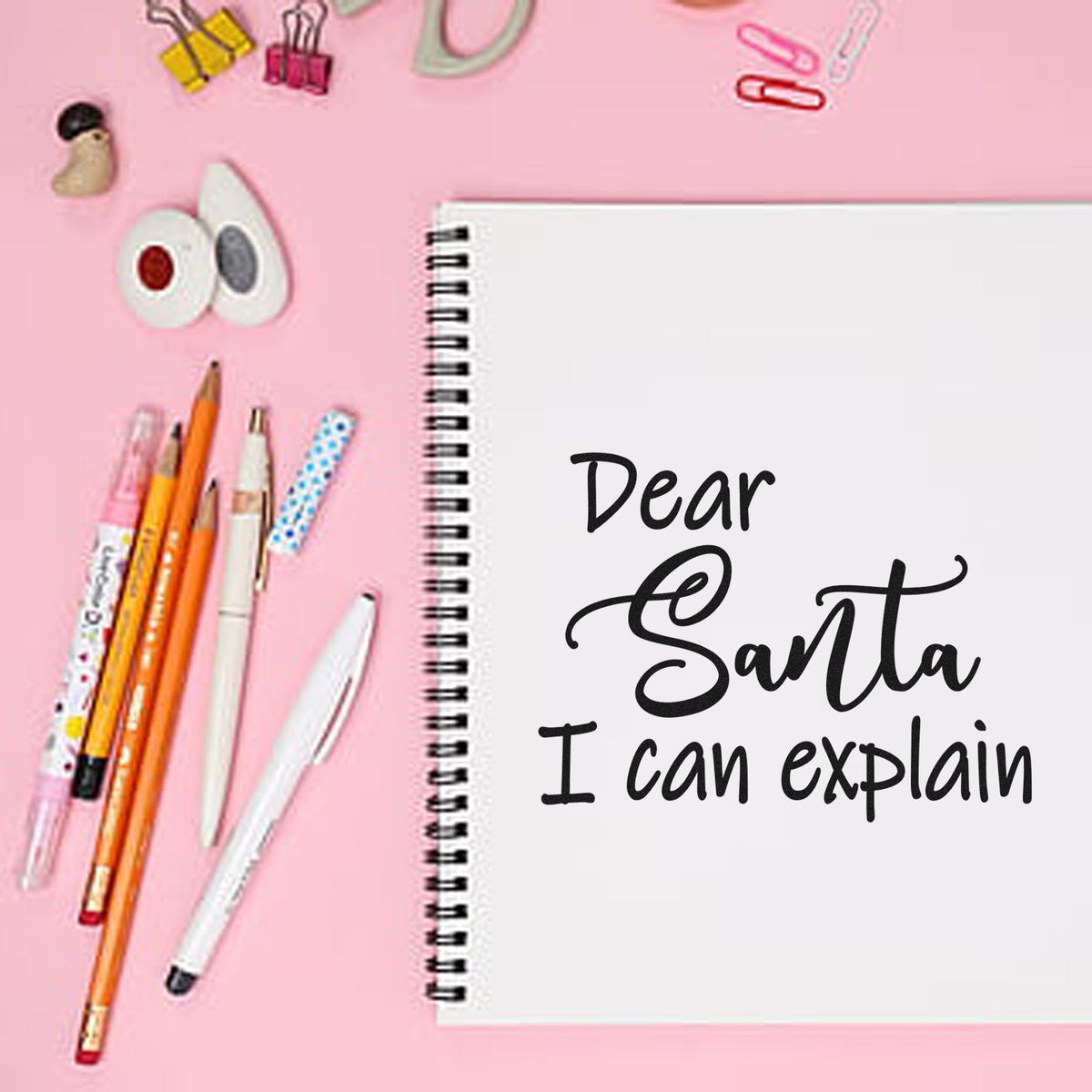 Pink stationery flat lay with stamp and seal products, spiral notebook reading Dear Santa I can explain, pens and clips.